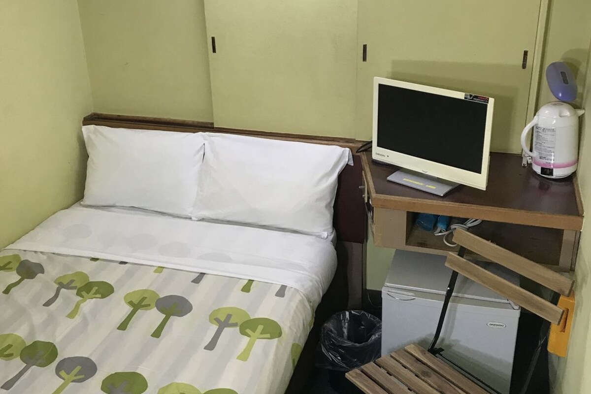 A small hotel room with a bed, a desk with a computer, a kettle, and a trash bin.