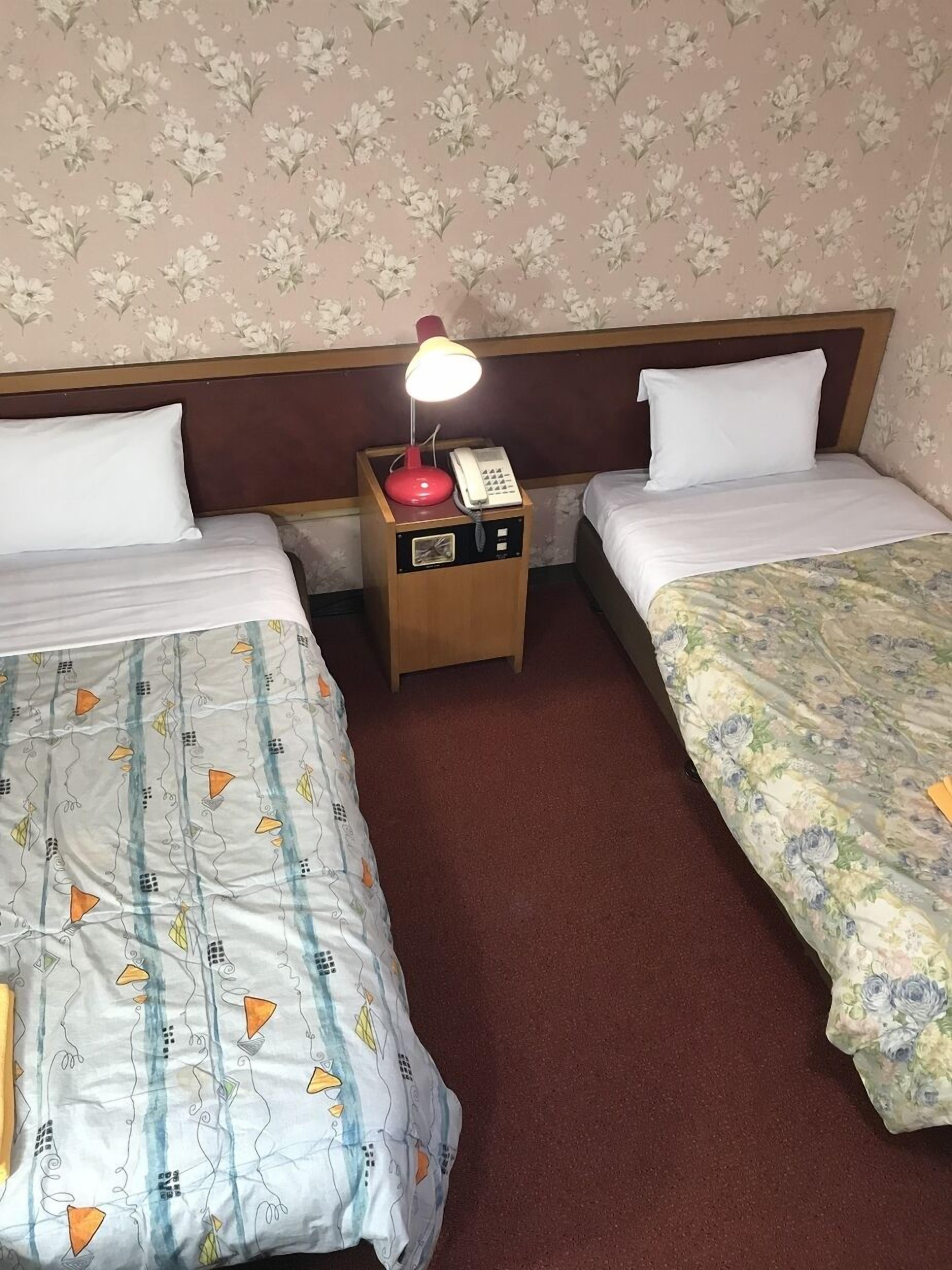Habitación de hotel con dos camas, una mesita de noche con lámpara y teléfono, papel tapiz con motivos florales y piso alfombrado.