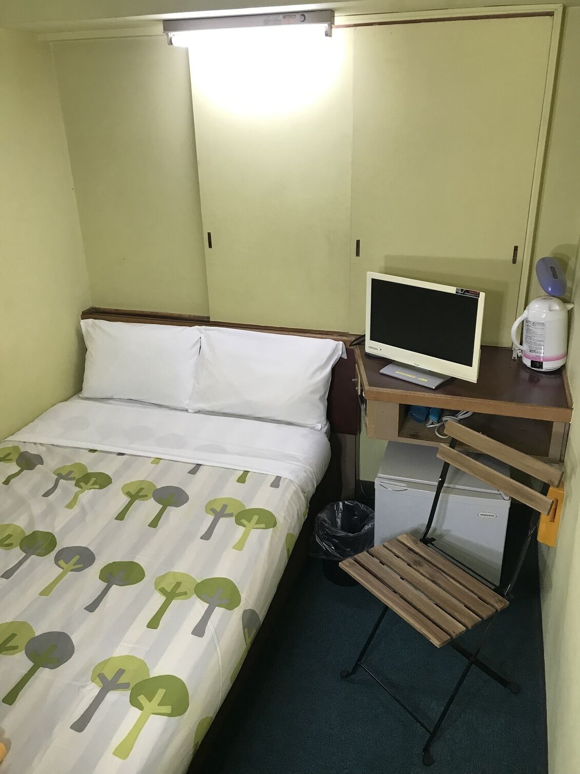 Una habitación de hotel pequeña con una cama, un escritorio con un ordenador, una tetera y un cubo de basura.