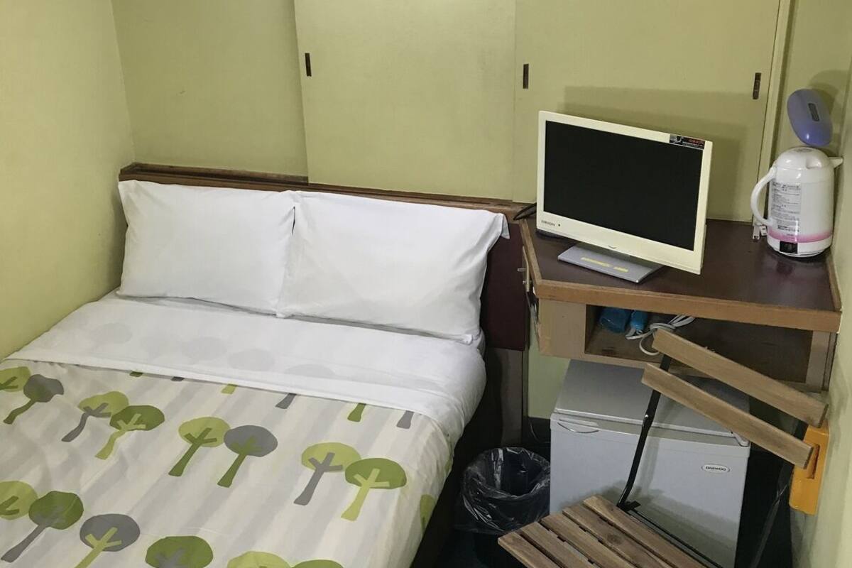 A small hotel room with a bed, a desk with a computer, a kettle, and a trash bin.
