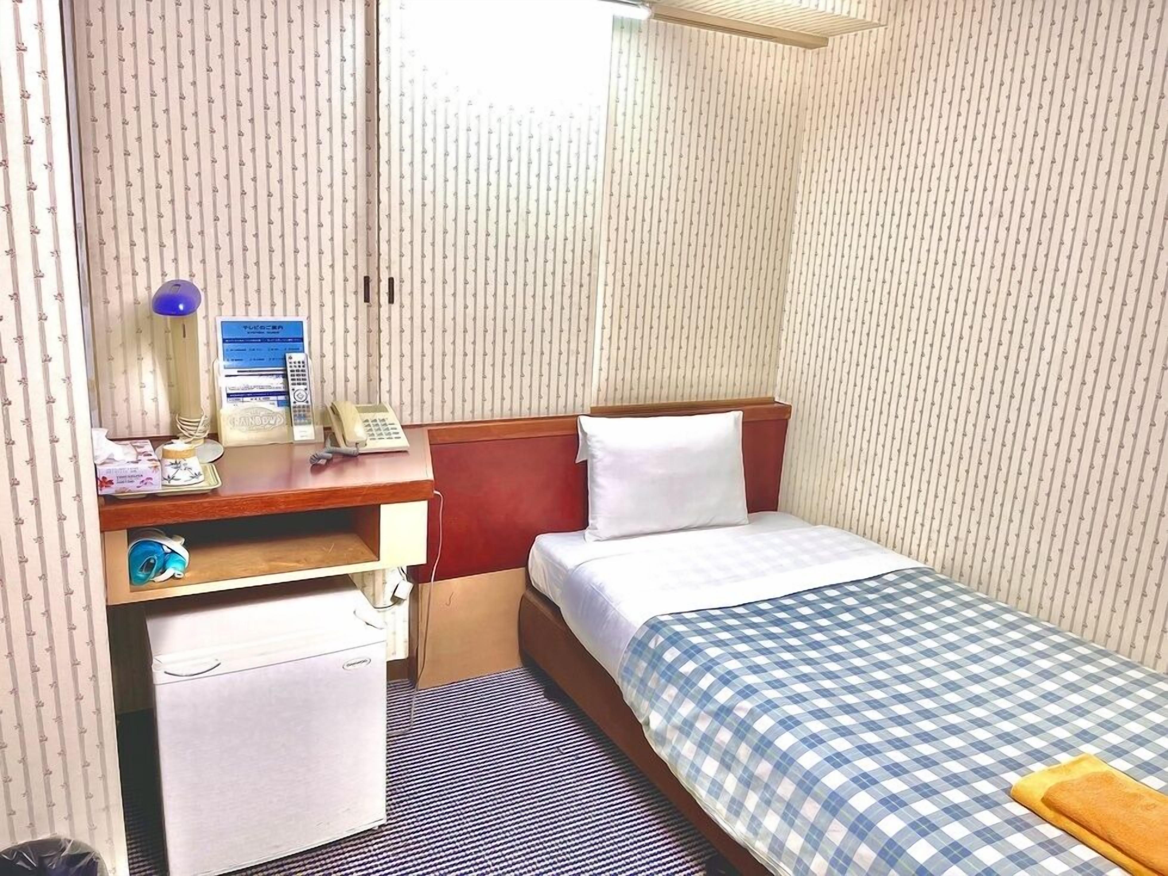 Habitación de hotel compacta con una cama individual, un escritorio con teléfono, un pequeño armario y una ventana con persianas verticales.