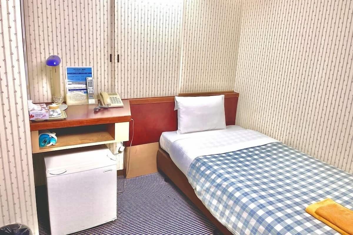 A compact hotel room with a single bed, a desk with a telephone, a small storage unit, and a window with vertical blinds.
