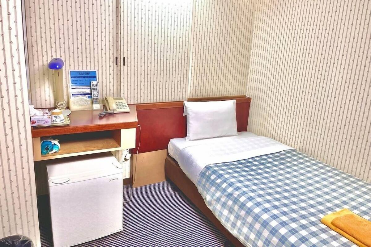 Habitación de hotel compacta con una cama individual, un escritorio con teléfono, un pequeño armario y una ventana con persianas verticales.