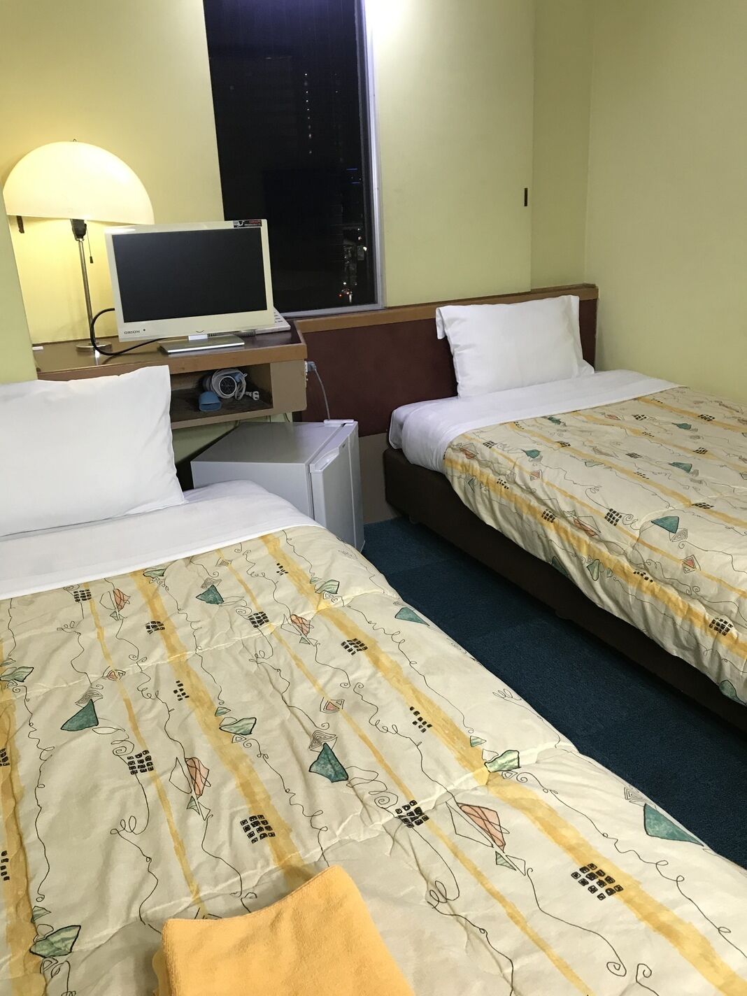 Una habitación de hotel con dos camas, un escritorio con ordenador, una lámpara y una ventana.