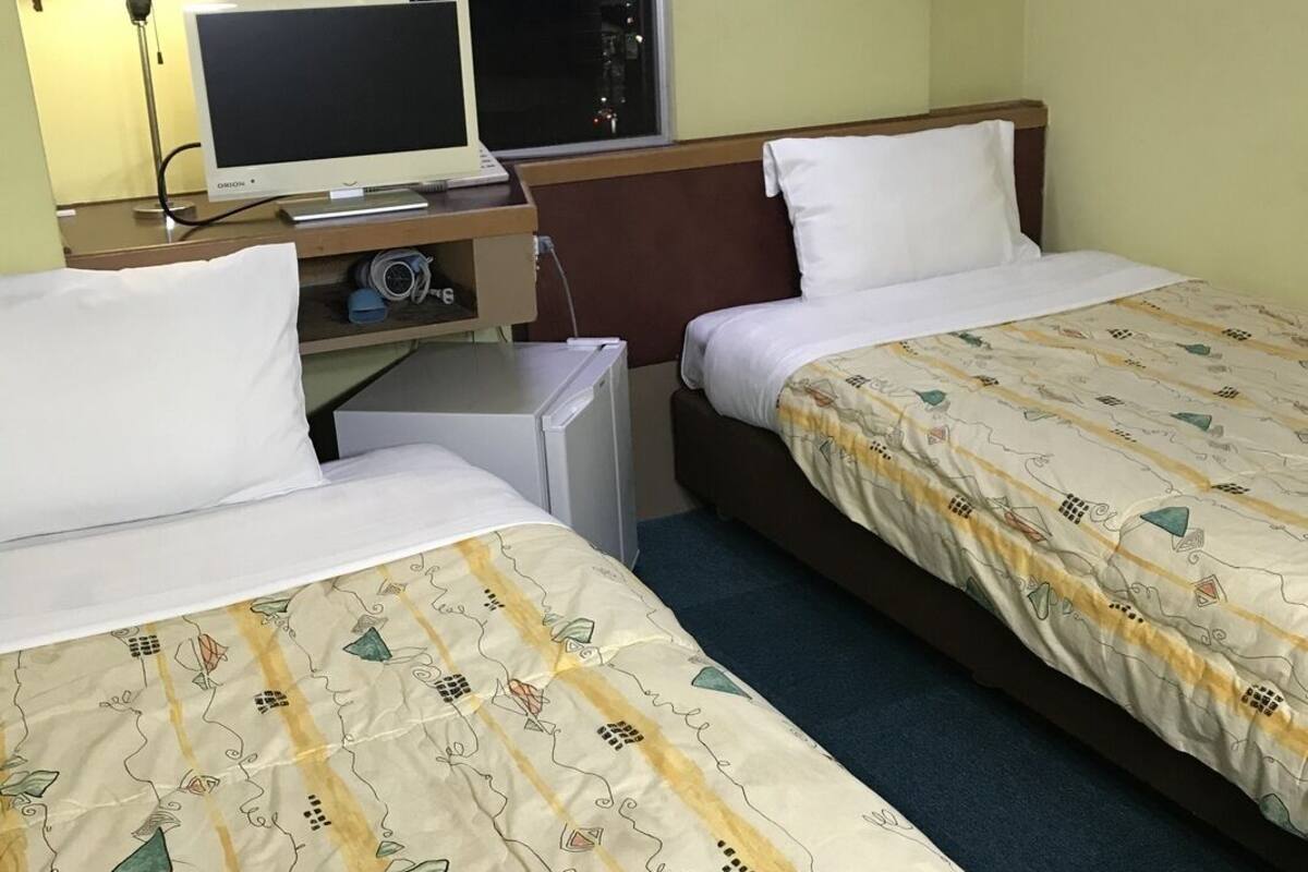 Una habitación de hotel con dos camas, un escritorio con ordenador, una lámpara y una ventana.