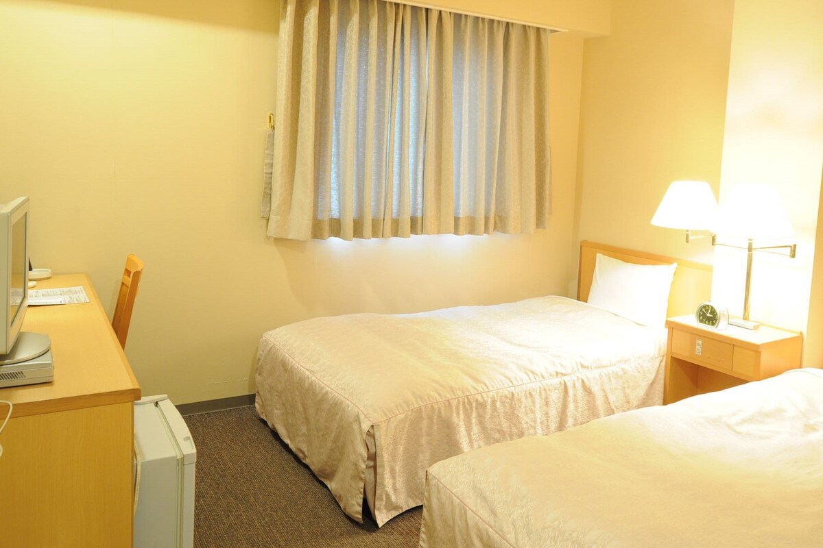 A hotel room with two beds, a desk with a computer, a lamp, and a window with curtains.