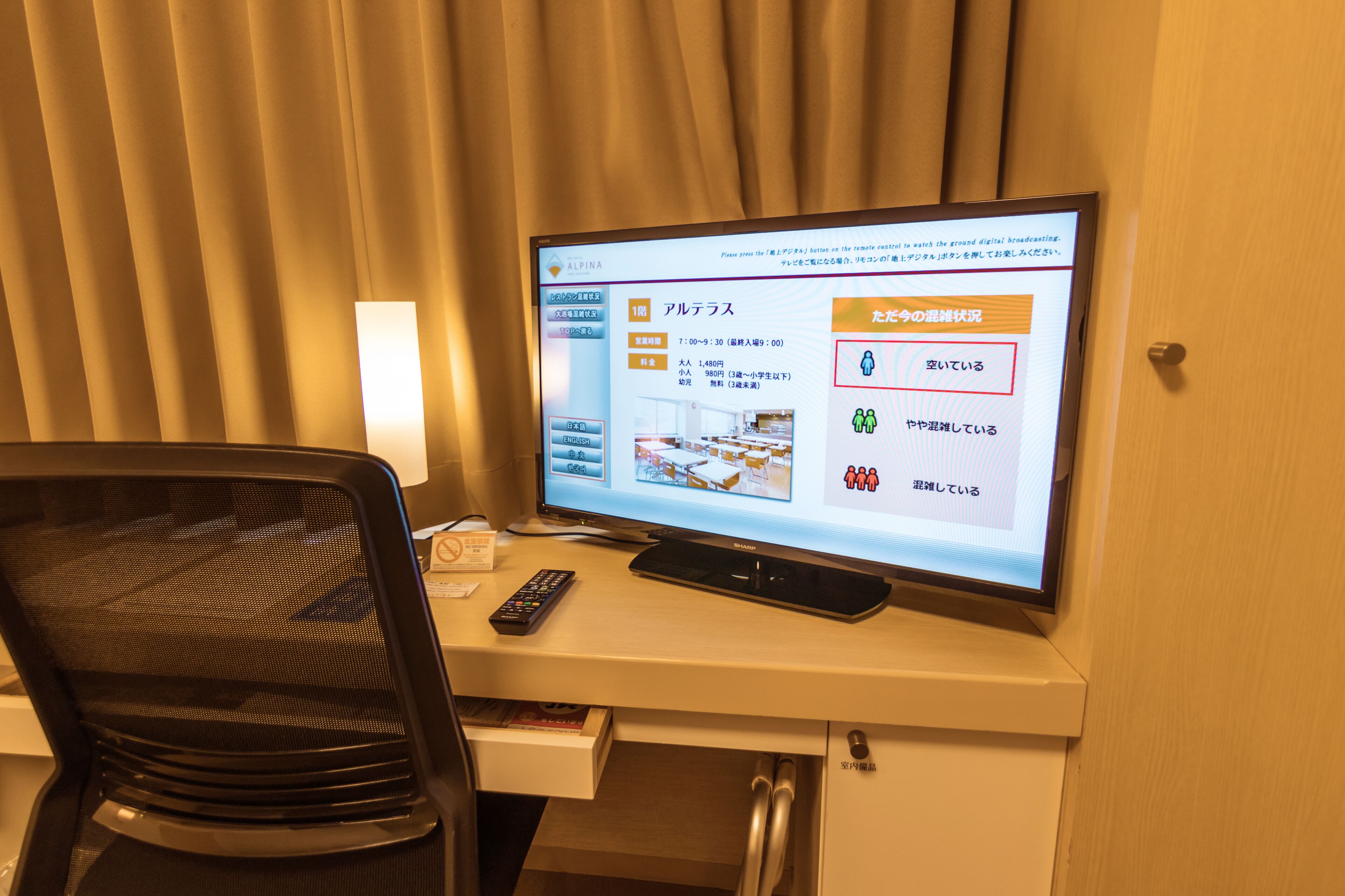 A hotel room with a desk, a computer monitor displaying a booking interface, a chair, and a small cabinet.