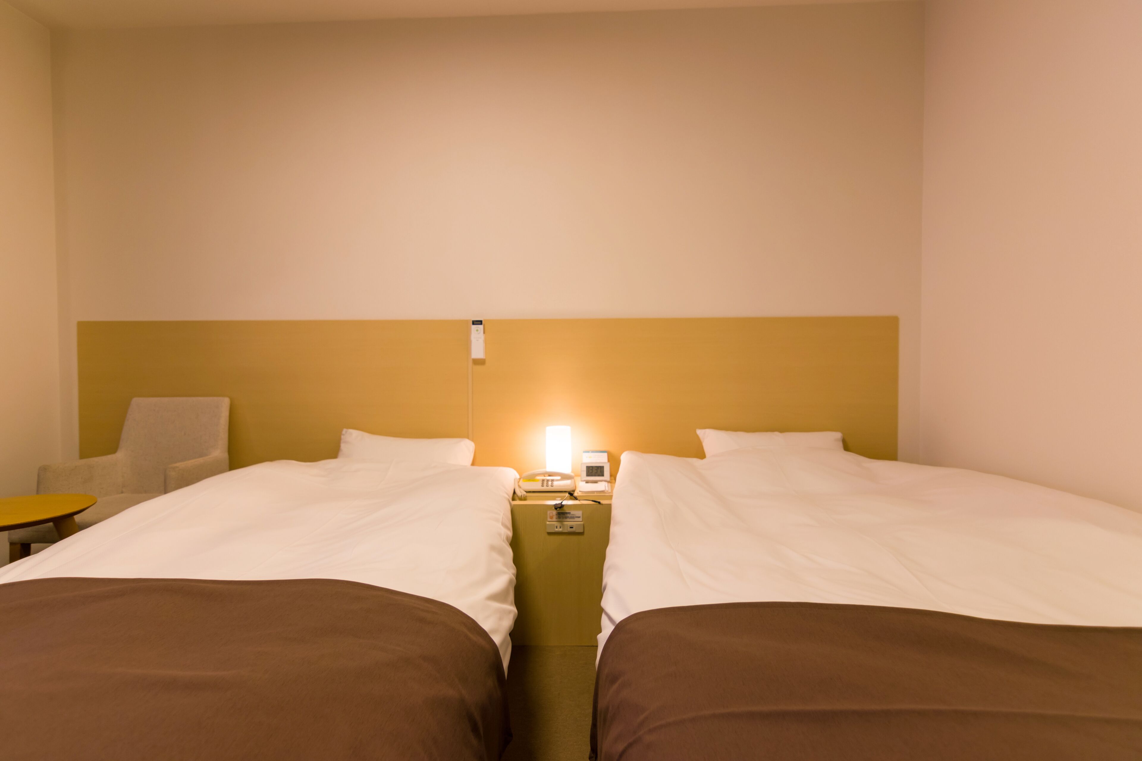 Habitación de hotel con dos camas, una mesita de noche con teléfono y lámpara, una silla y una pequeña mesa.