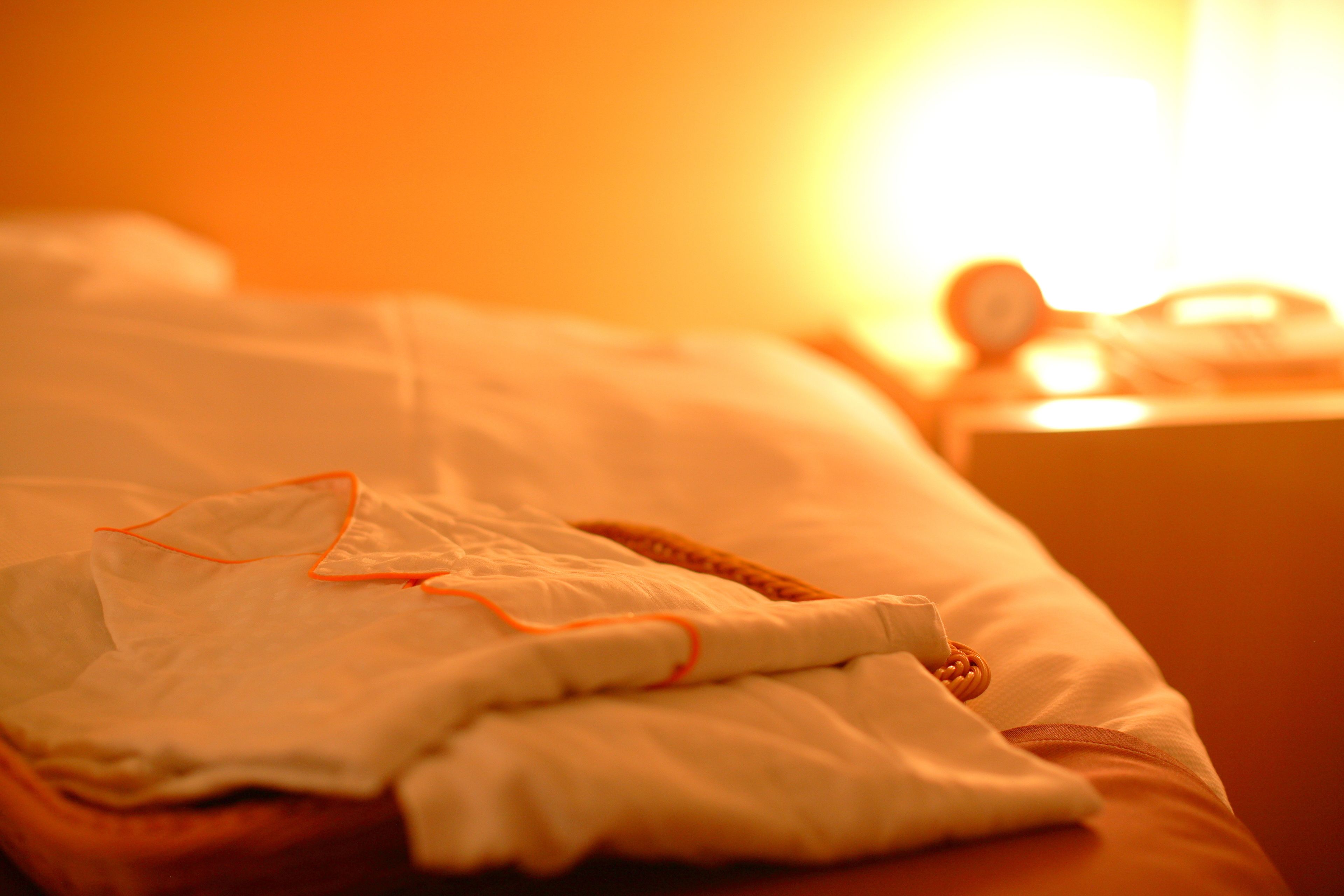 Una cama con una manta y almohada blancas, una mesita de noche con un reloj, y una fuente de luz cálida al fondo.