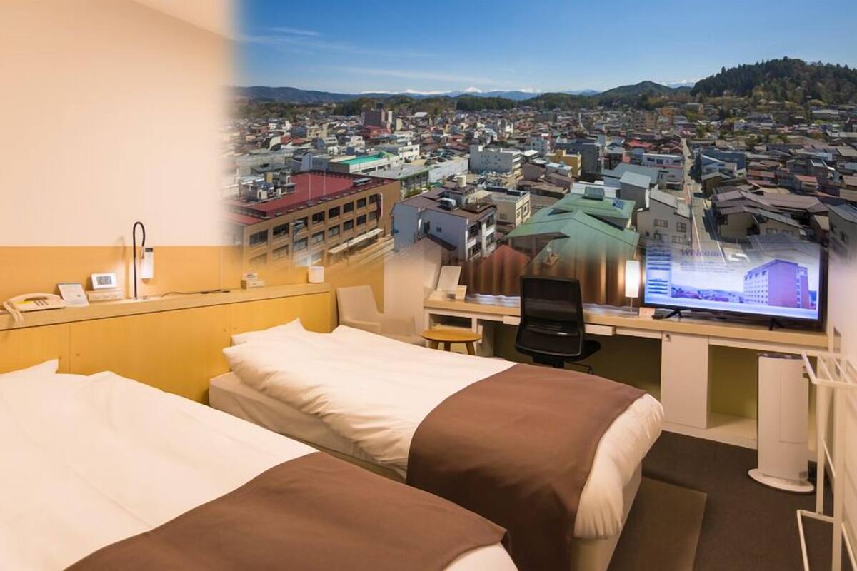 A hotel room with two beds, a desk with a computer, and a view of a cityscape.