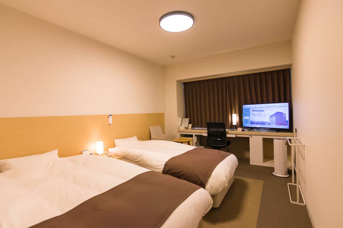 A hotel room with two beds, a desk with a computer, a chair, and a television displaying a welcome screen.