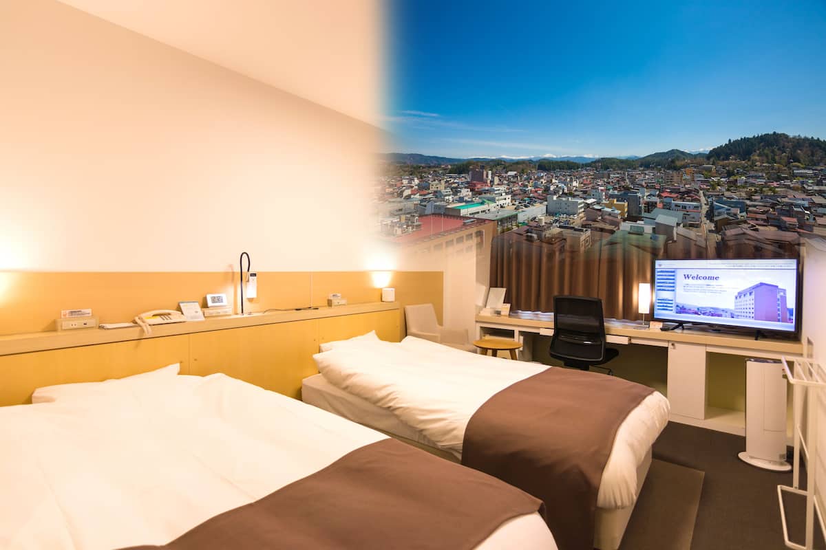 A hotel room with two beds, a desk with a computer, and a view of a cityscape.