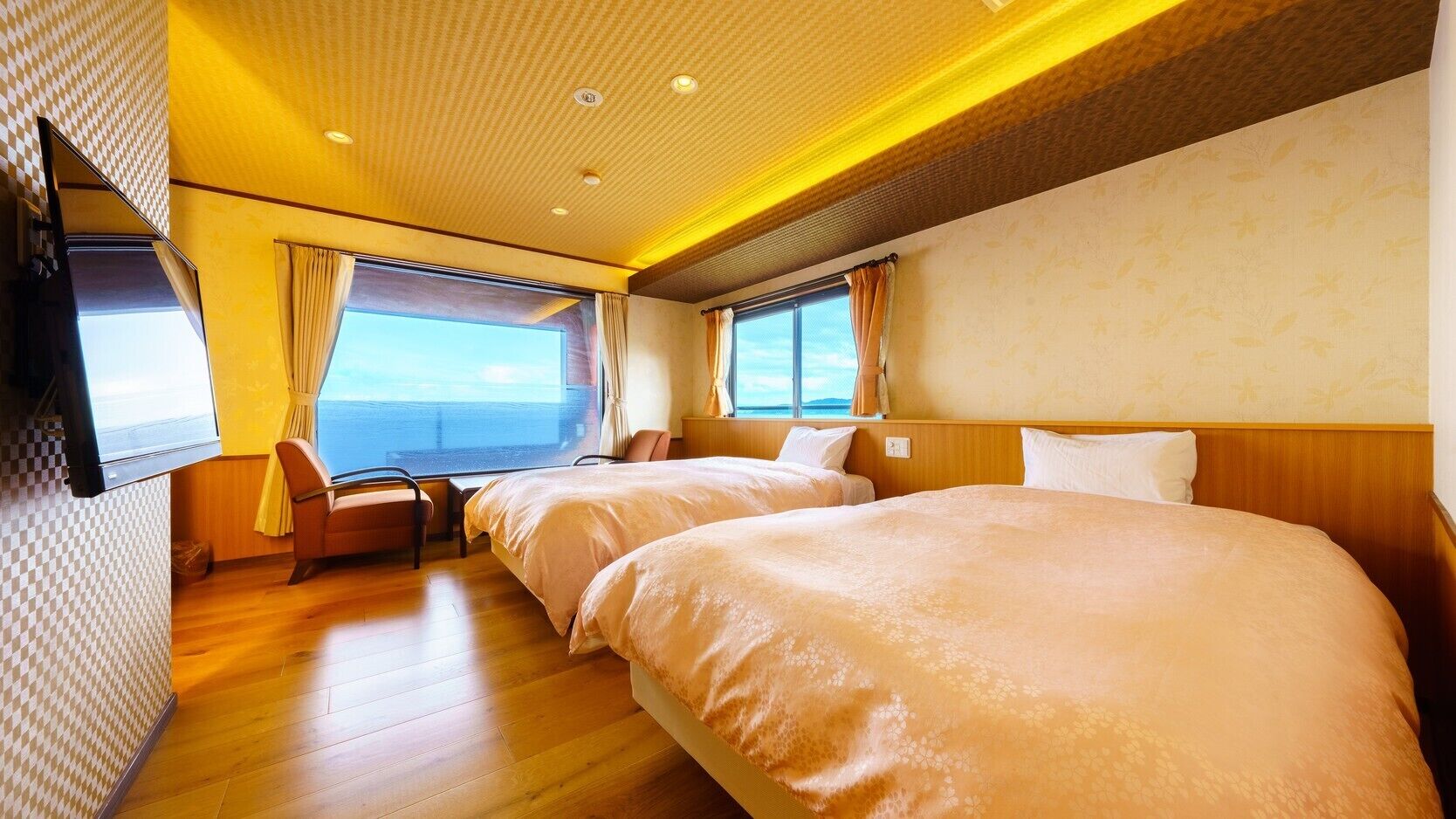 Une chambre d’hôtel avec deux lits, un coin salon avec un fauteuil, une télévision et de grandes fenêtres donnant sur la mer.