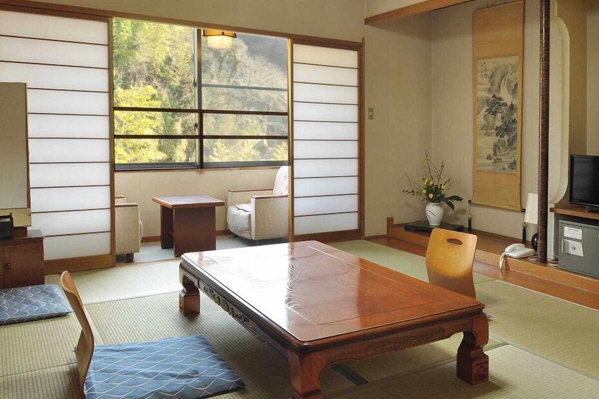 A traditional Japanese room with tatami flooring, a low wooden table, a sofa, a chair, a small side table, a TV, and a painting on the wall.