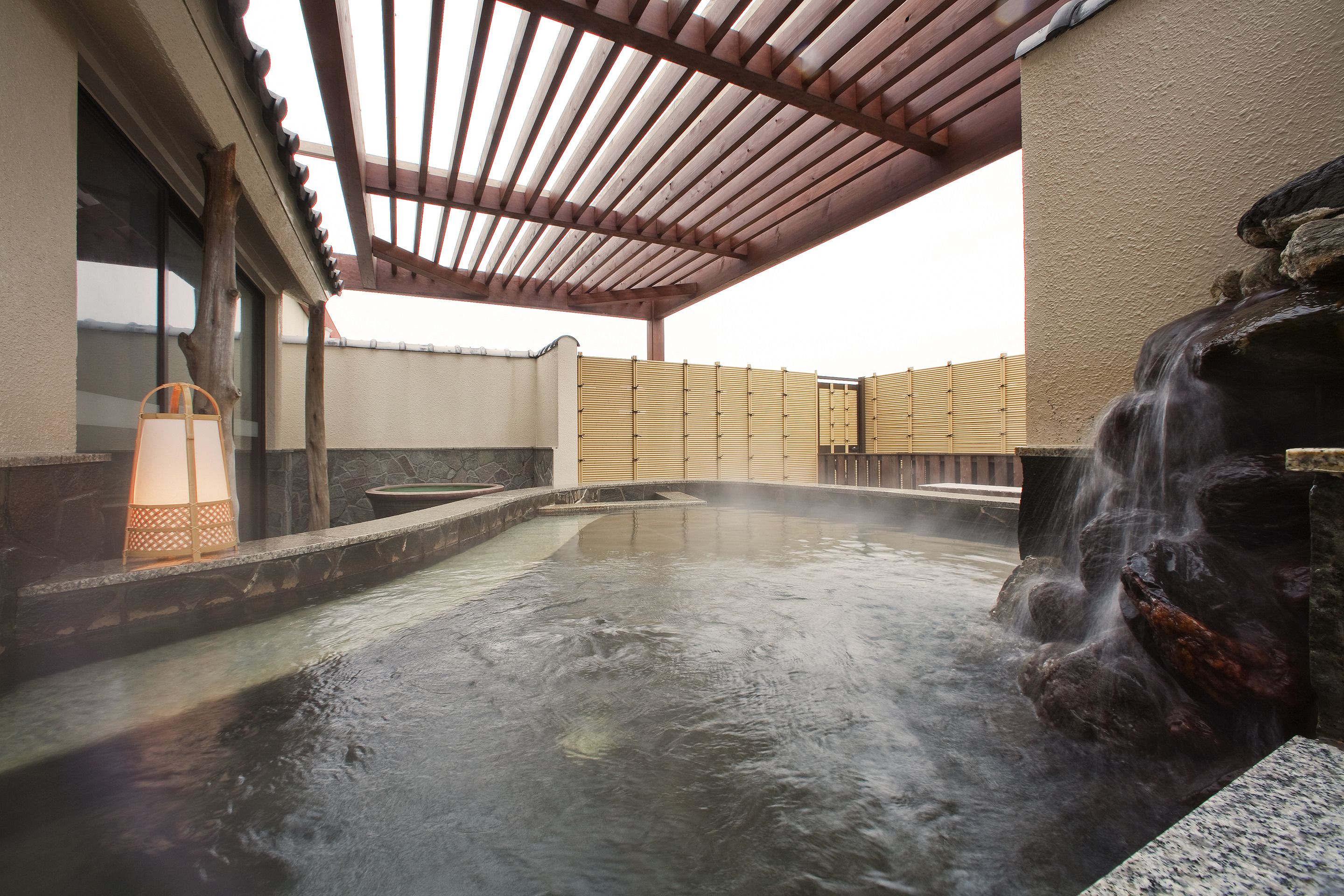 A hot spring area with a wooden pergola, stone walls, and a waterfall feature.