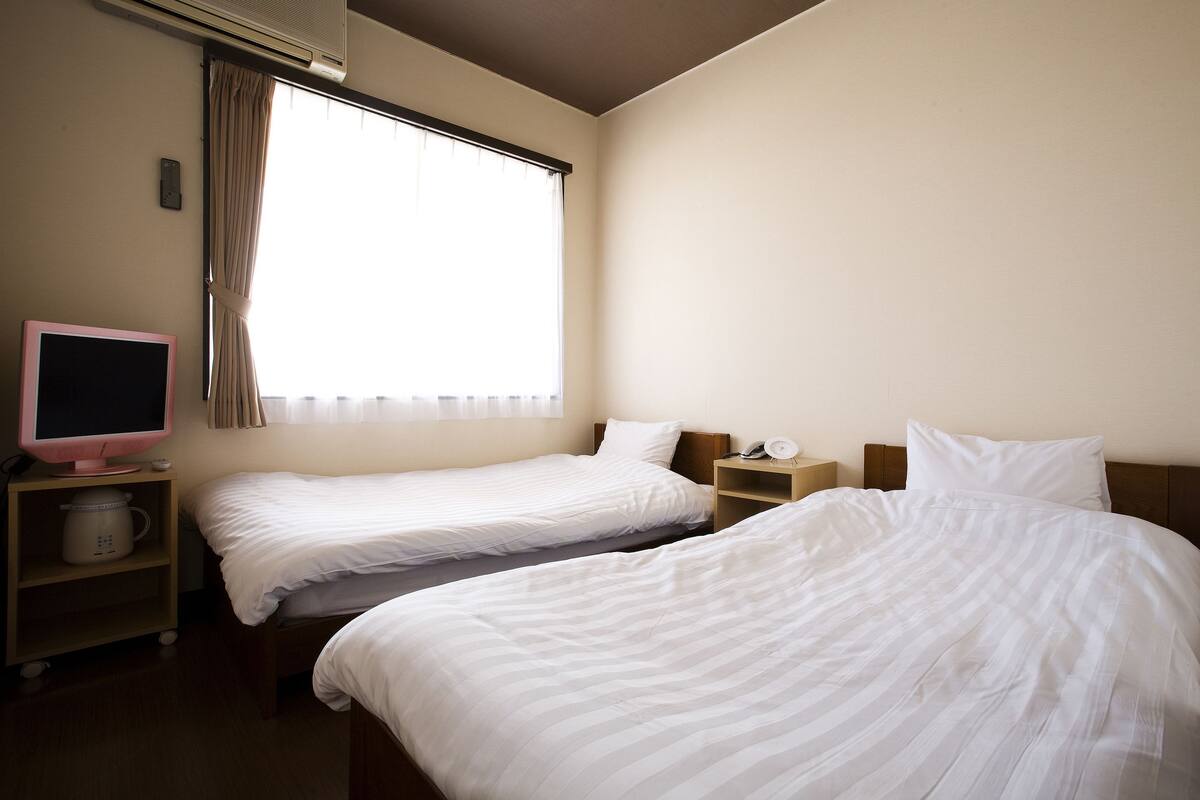 A hotel room with two single beds, a bedside table with a lamp, a television, and a shelf with a kettle.