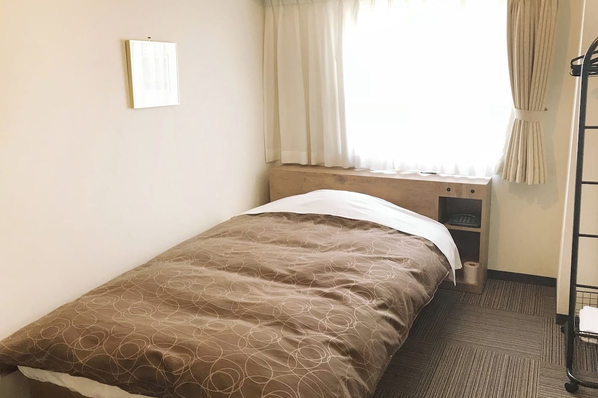 Una habitación pequeña con cama individual, ventana, cortina, mesita de noche y estante fijado a la pared.