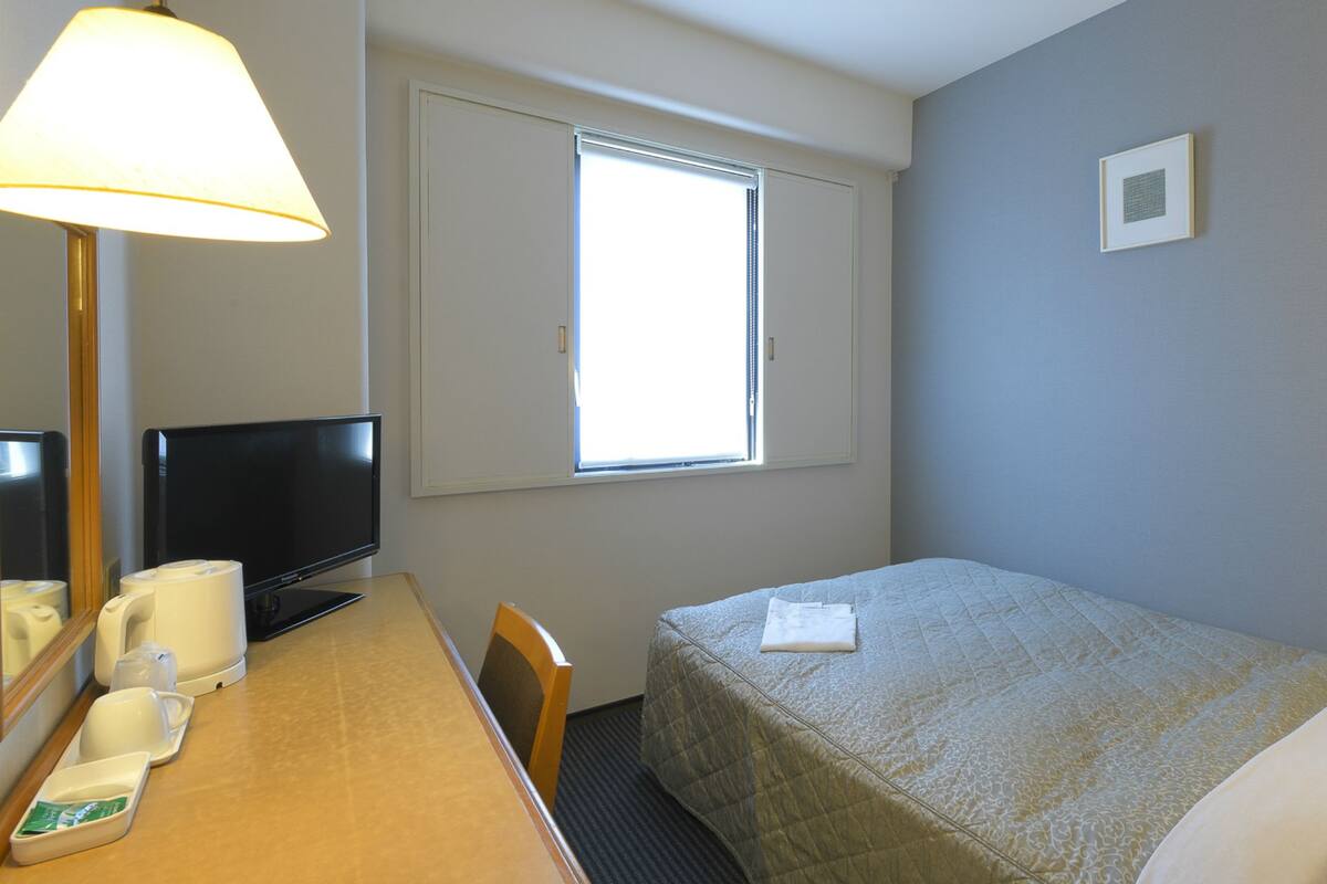 A hotel room with a bed, a desk with a telephone, a chair, a television, and a window with blinds.