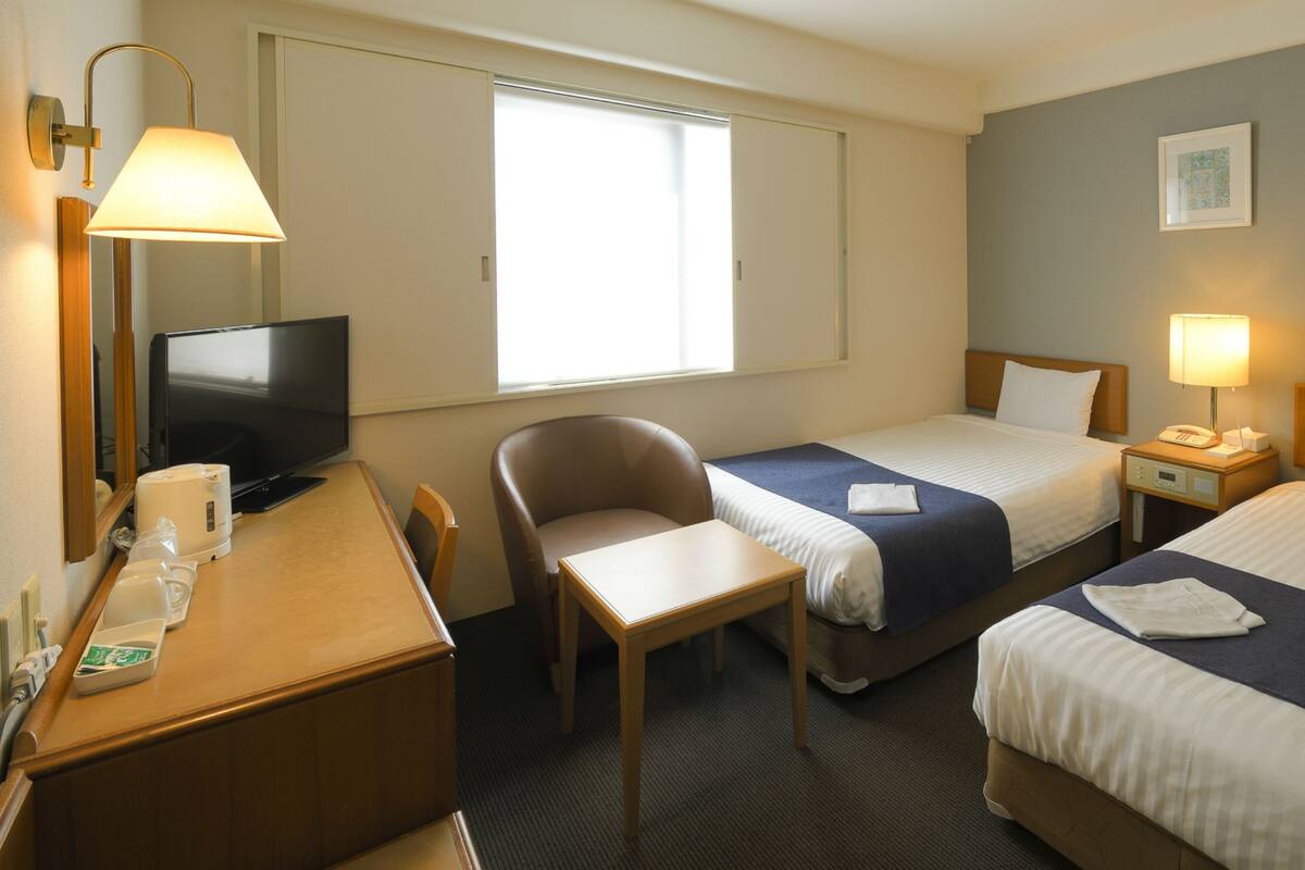 A hotel room with two beds, a desk, a chair, a television, and a window.