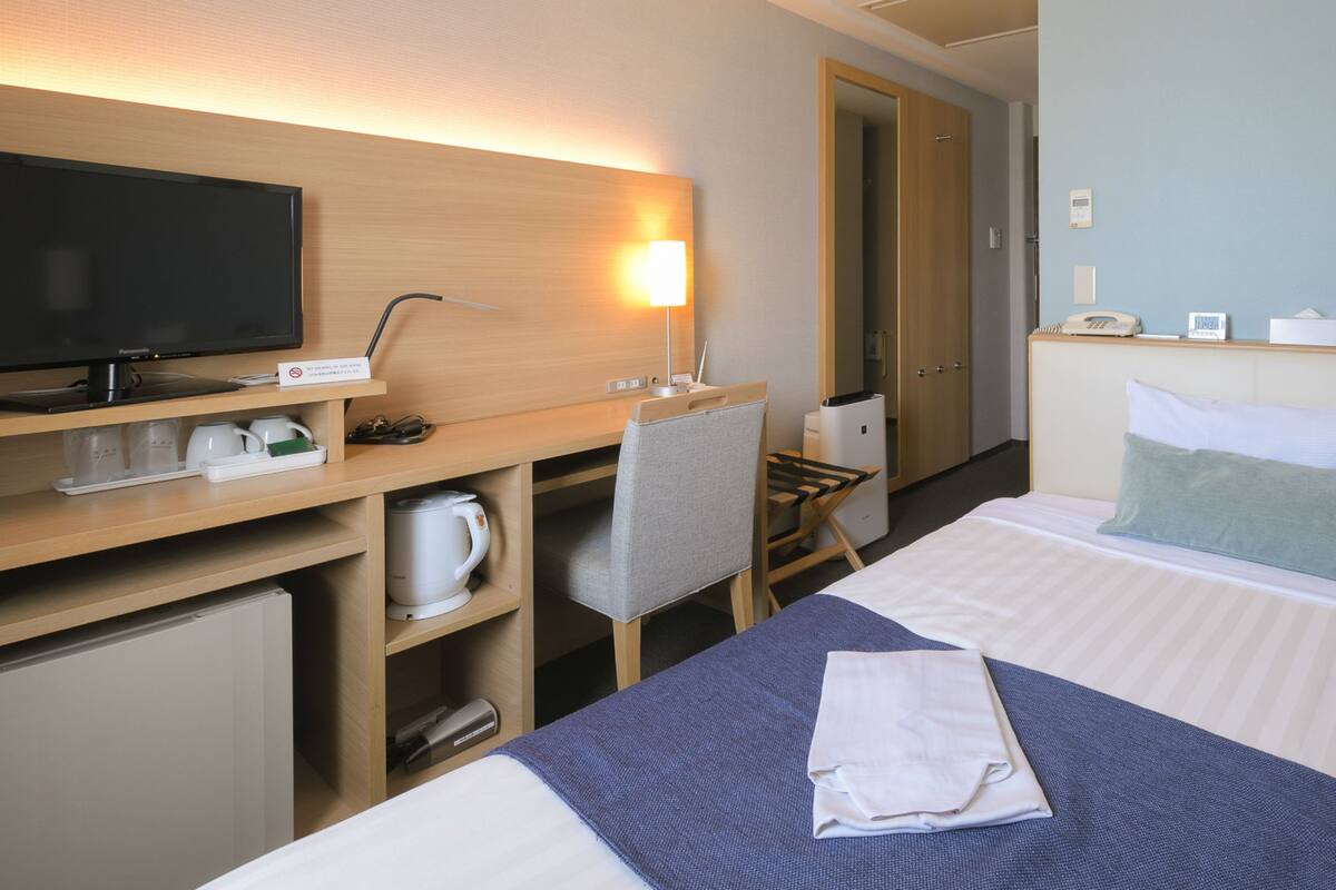 A hotel room with a bed, a desk with a computer and a lamp, a TV, and a small kitchenette.