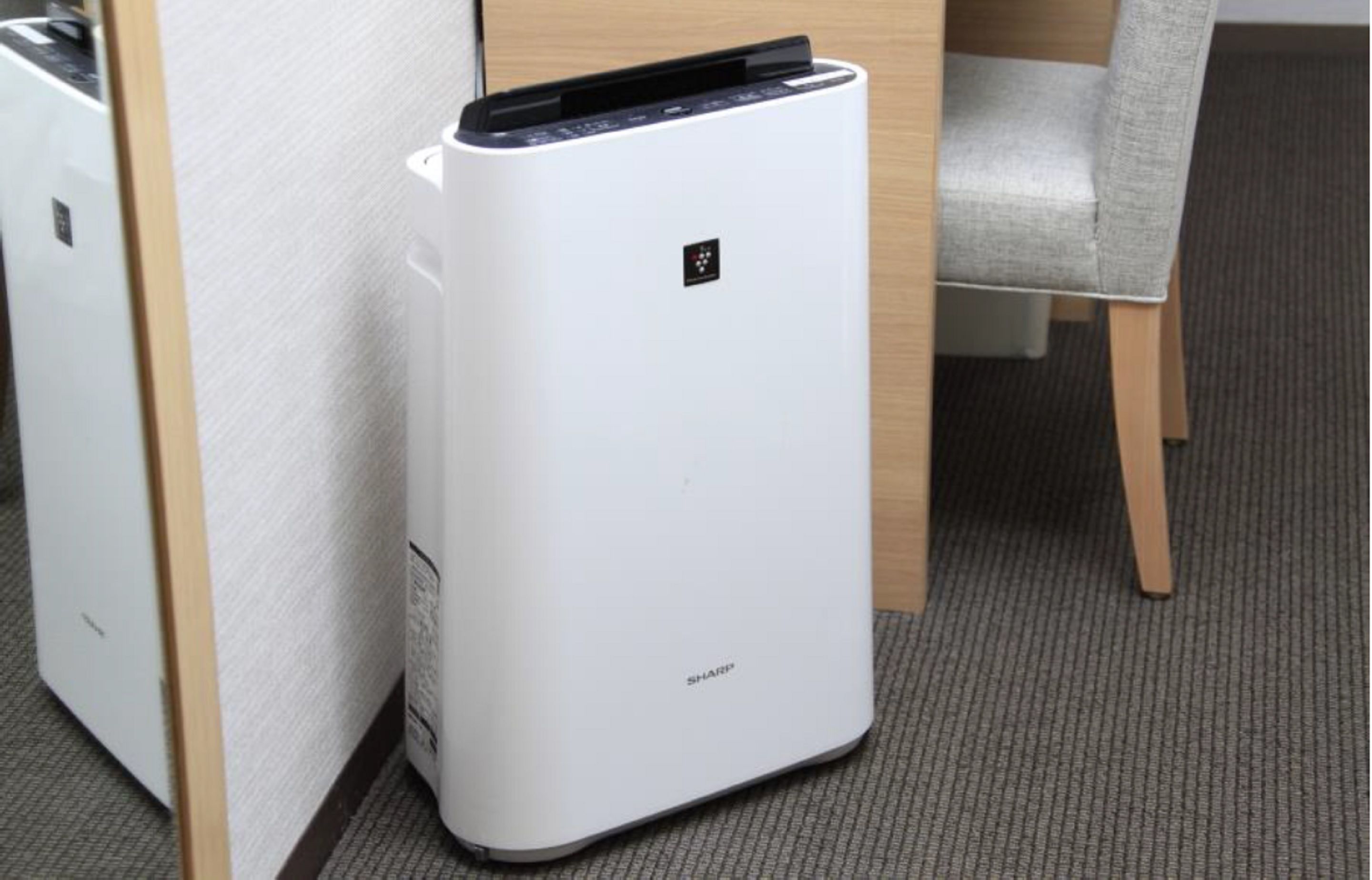 Purificador de aire blanco con panel de control, ubicado sobre una alfombra cerca de un escritorio de madera y una silla gris.