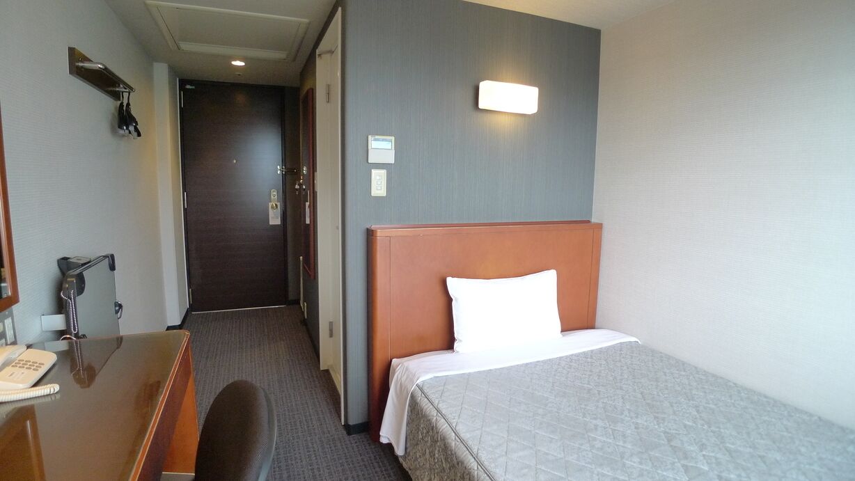 Ein Hotelzimmer mit einem Bett, einem Schreibtisch mit Telefon, einem Stuhl, einer Tür und einer Wandleuchte.