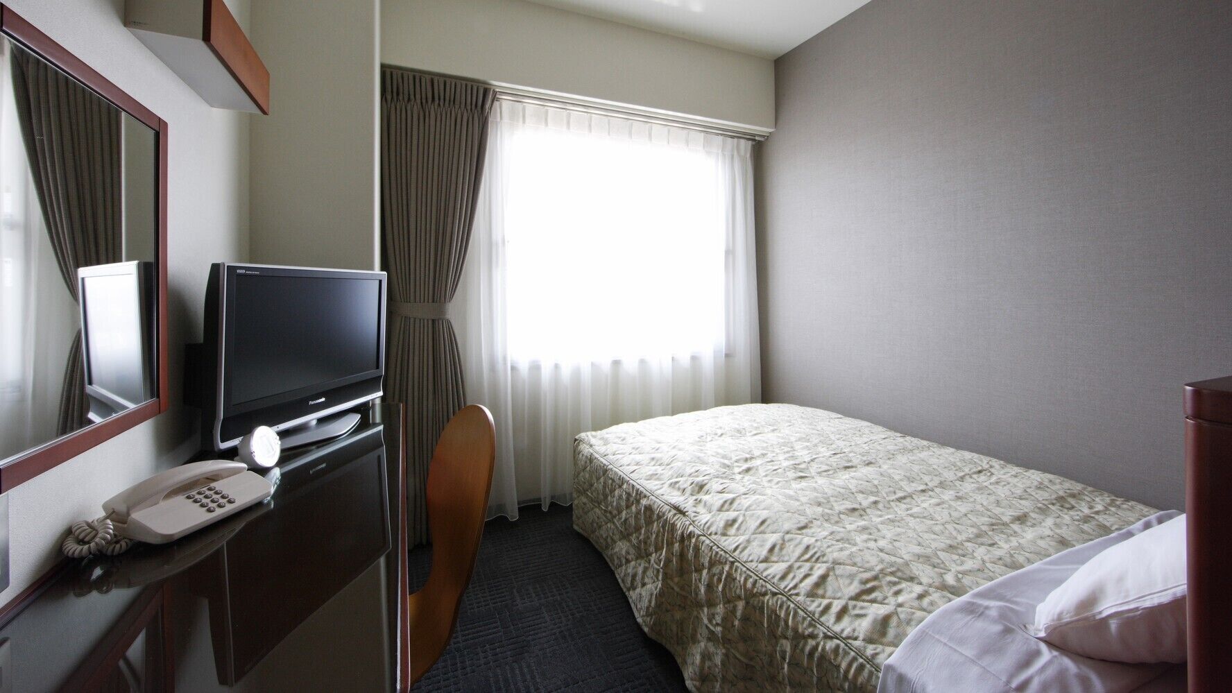 Ein Hotelzimmer mit einem Bett, einem Schreibtisch mit Telefon und Fernseher, einem Sessel und einem Fenster mit Vorhängen.