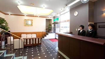 A hotel reception area with a reception desk, two receptionists, a staircase, and a large window.