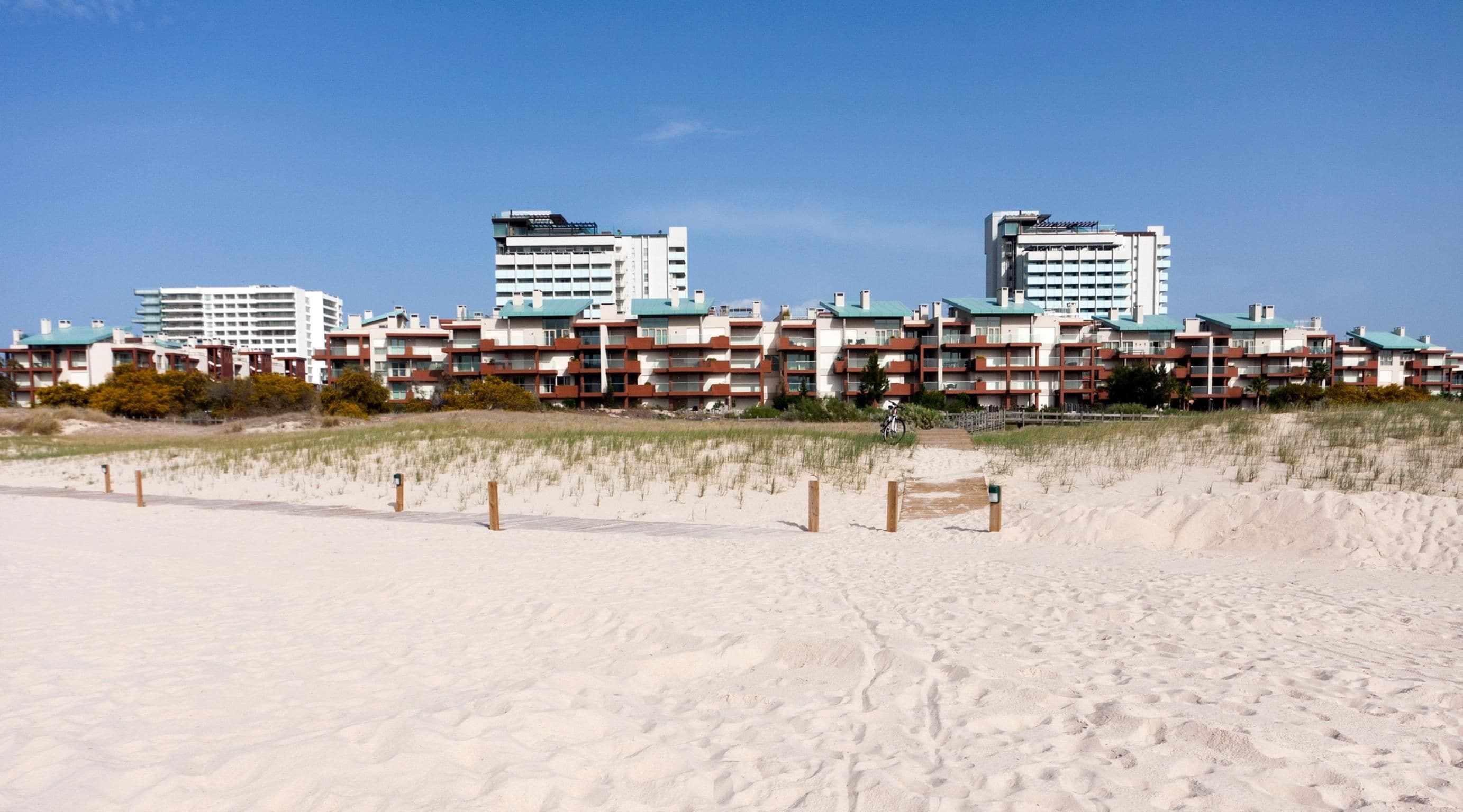 A beachfront hotel with multiple floors, a sandy beach in the foreground, and a clear sky.