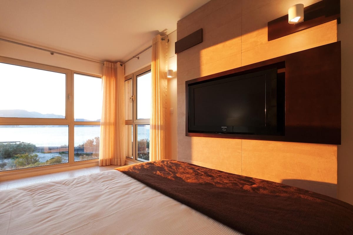 Habitación de hotel con una cama grande, un televisor de pantalla plana montado en la pared y vistas al mar a través de amplios ventanales.
