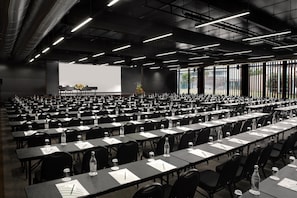 Un salón de conferencias con filas de mesas, sillas y una pantalla grande para presentaciones.