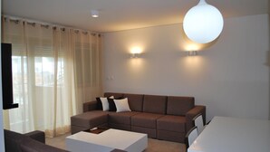 Una sala moderna con un sofá modular, una mesa de centro blanca y una mesa de comedor. Hay dos luces empotradas en la pared y una lámpara en el techo.