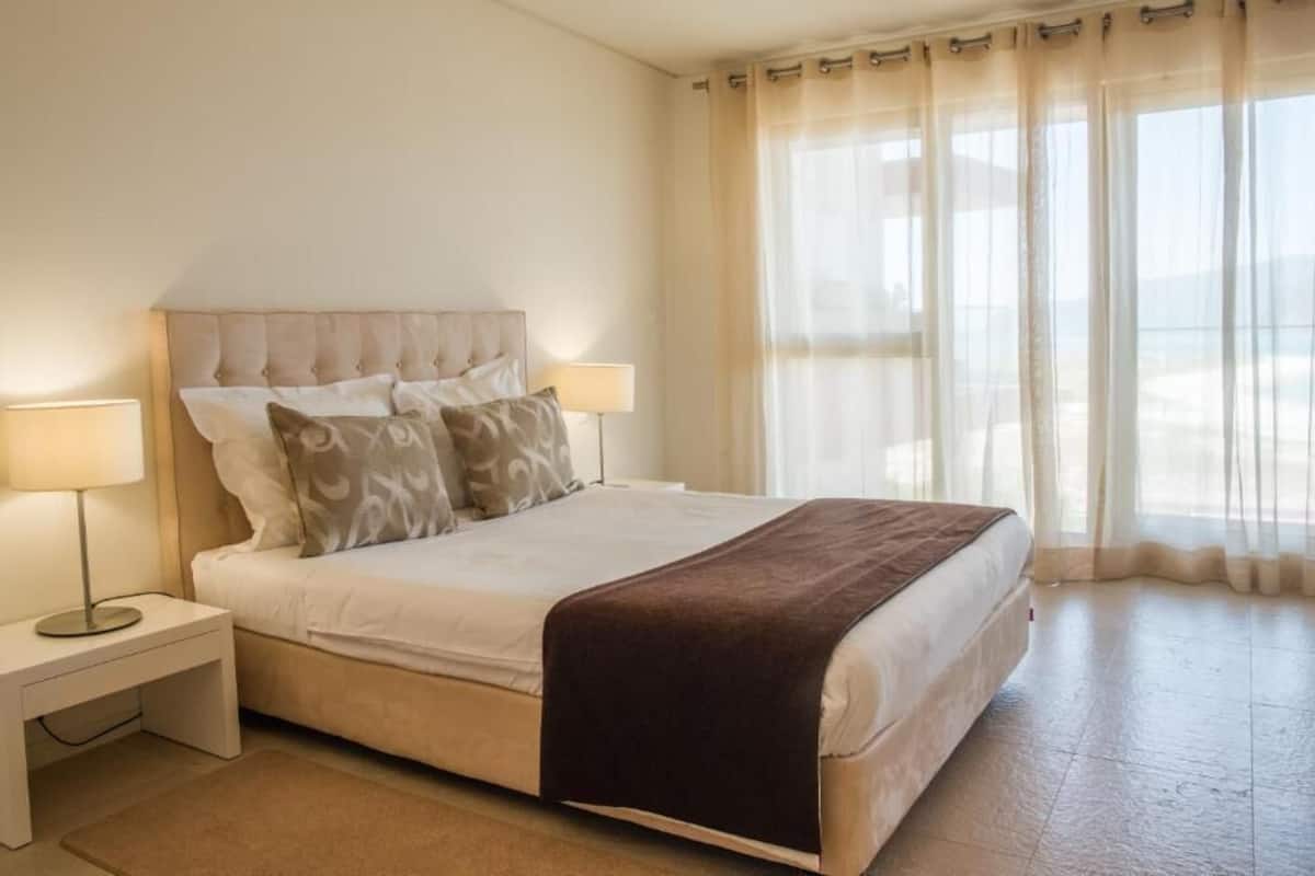 Una cama bien hecha con una manta marrón, dos lámparas de noche y una ventana con cortinas translúcidas.