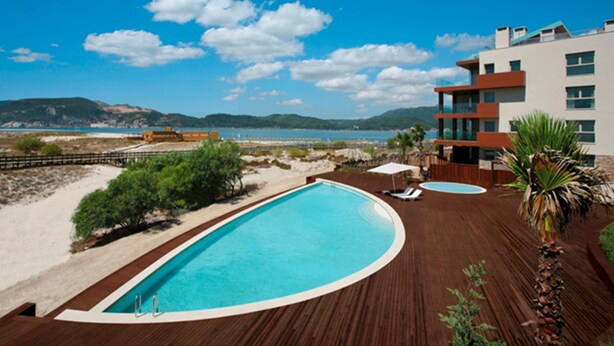Una piscina con boscaglia, spiaggia e un edificio moderno con balconi.