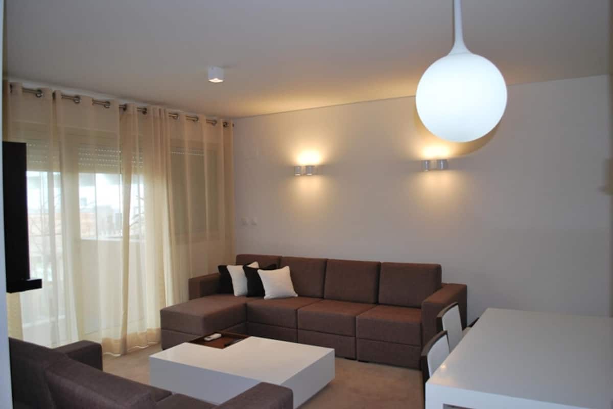 Una sala moderna con un sofá seccional, una mesa de centro blanca y una mesa de comedor. Hay dos luces empotradas en la pared y una lámpara de techo.