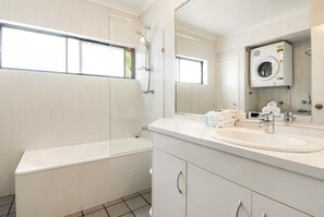 A modern bathroom with a bathtub, a sink, a mirror, and a built-in washing machine. - Cape Apartments (Byron Bay)