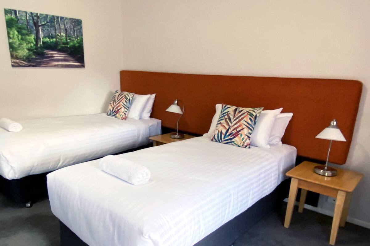 A hotel room with two single beds, each with a pillow and a rolled towel. There are two bedside tables with lamps.