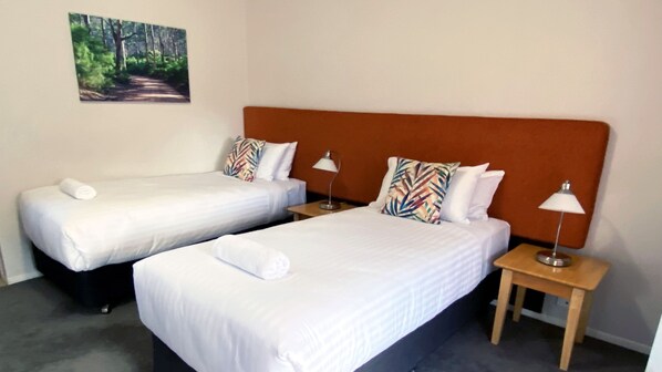 Habitación de hotel con dos camas individuales, cada una con una almohada y una toalla enrollada. Hay dos mesitas de noche con lámparas.