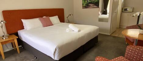 Une chambre d’hôtel avec un grand lit, une table de chevet avec une lampe, une chaise et une petite table sur laquelle est posé un vase de fleurs.