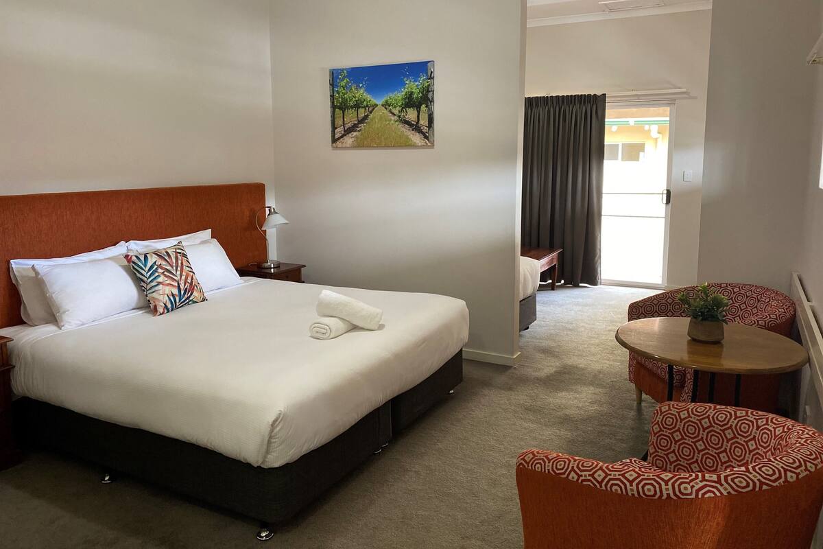 A hotel room with a large bed, a nightstand, a lamp, a painting on the wall, a view of the outside, and a small table with a plant.