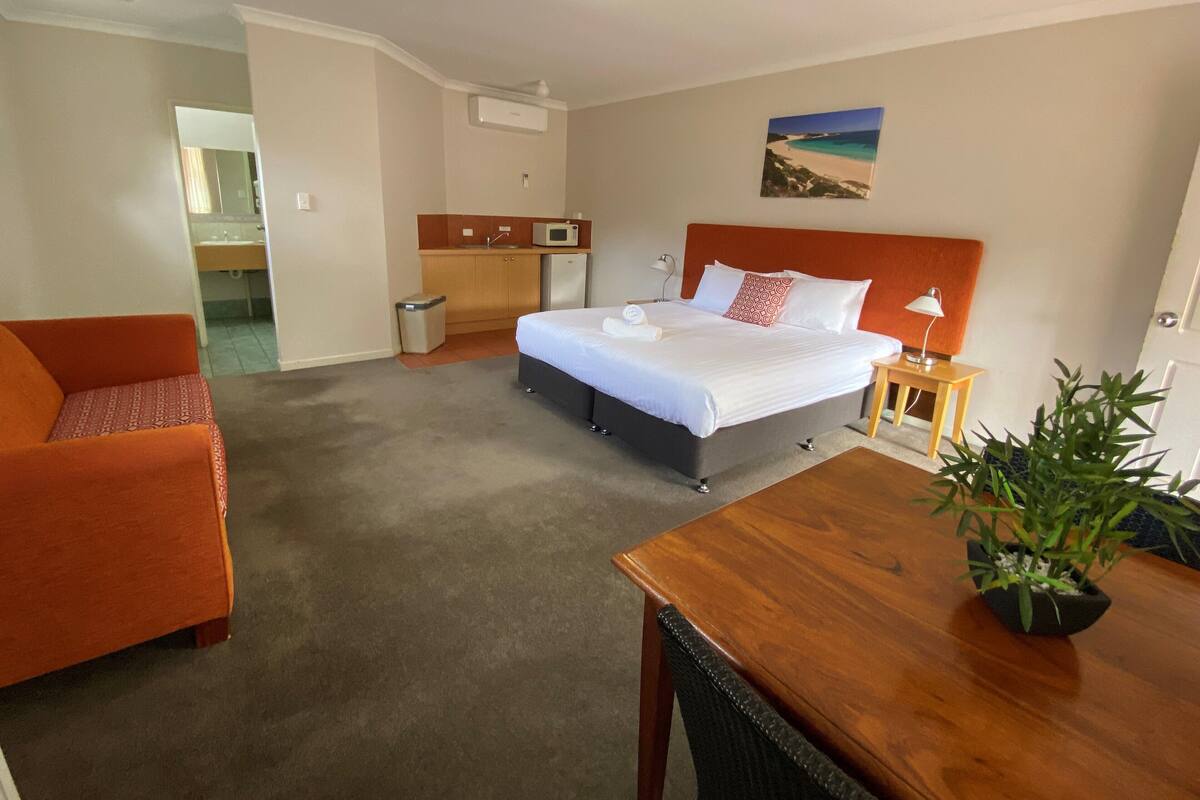 A hotel room with a bed, a desk, a chair, a small table, a trash bin, a microwave, a sink, and a potted plant.