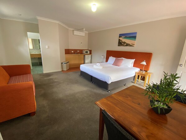 Habitación de hotel con una cama, un escritorio, una silla, una mesita, un cubo de basura, un microondas, un lavamanos y una planta en maceta.