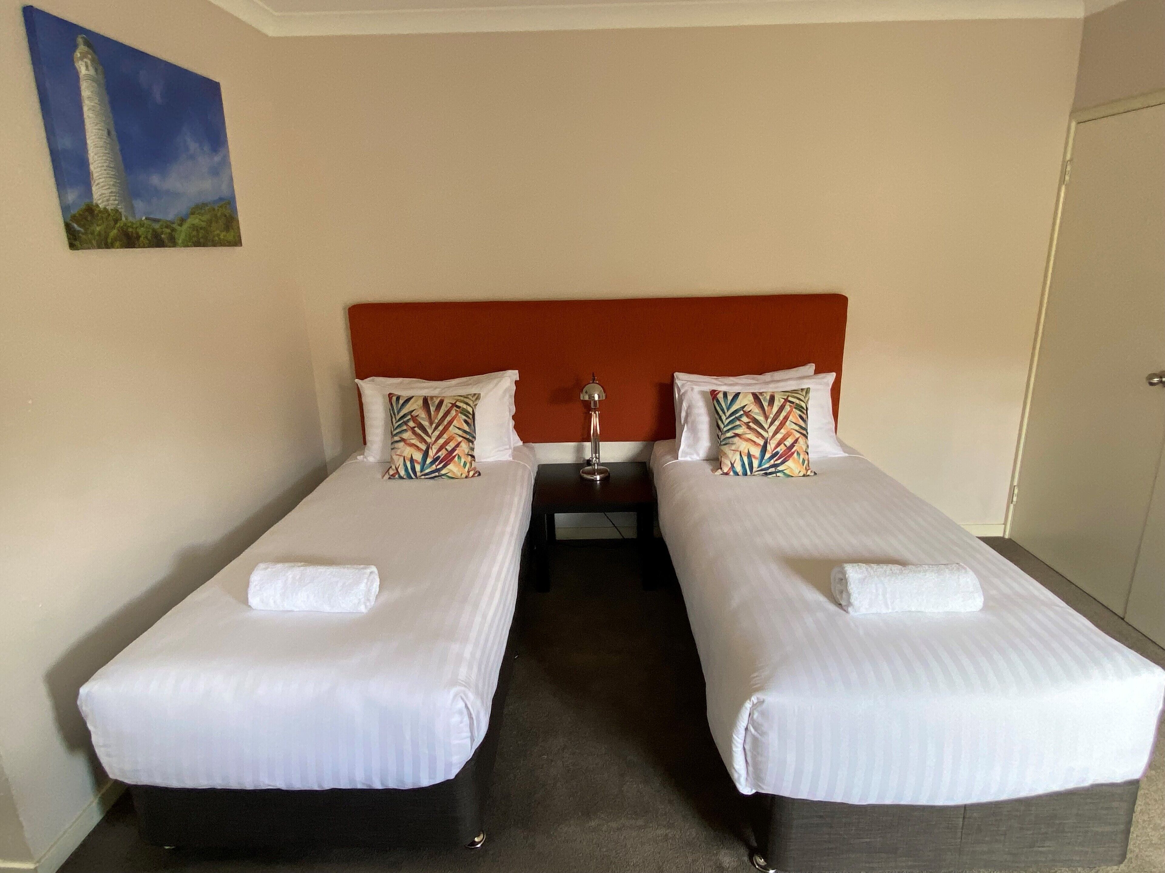 Habitación de hotel con dos camas individuales, cada una con una colcha blanca y almohadas a juego. Hay una mesita de noche con una lámpara entre las camas, y un cuadro de un faro en la pared.