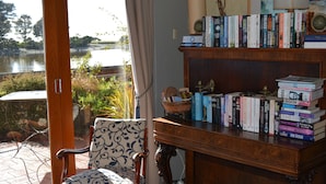 A room with a view of a body of water, a wooden bookshelf with books, a patterned chair, and a table with pens. - Burnie Airport Motel (Wynyard)