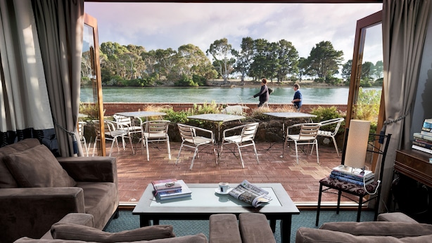 A living room with a view of a lake, outdoor seating, and a table with books and a cup. - Burnie Airport Motel (Wynyard)