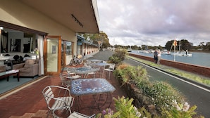 An outdoor cafe with tables and chairs, a bike parked by the entrance, and a view of a canal with boats. - Burnie Airport Motel (Wynyard)