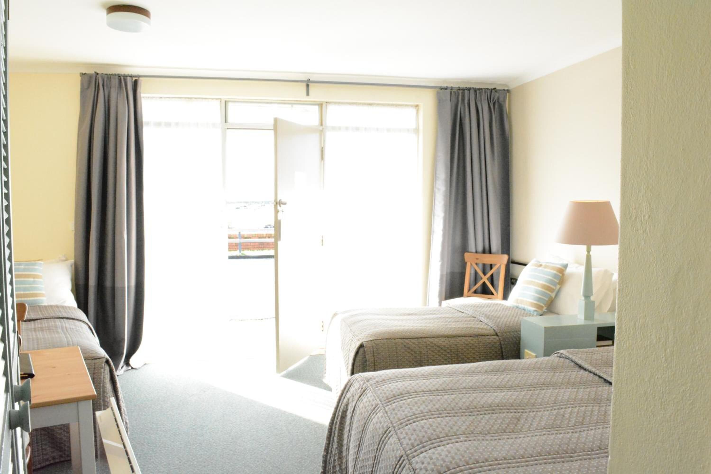 A hotel room with two beds, a small table, a lamp, and a view through a sliding door.