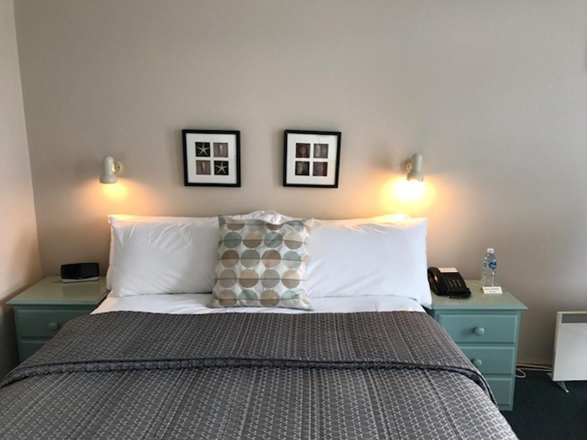 A hotel room with a bed, two bedside tables, a nightstand, a lamp, and two framed pictures on the wall.
