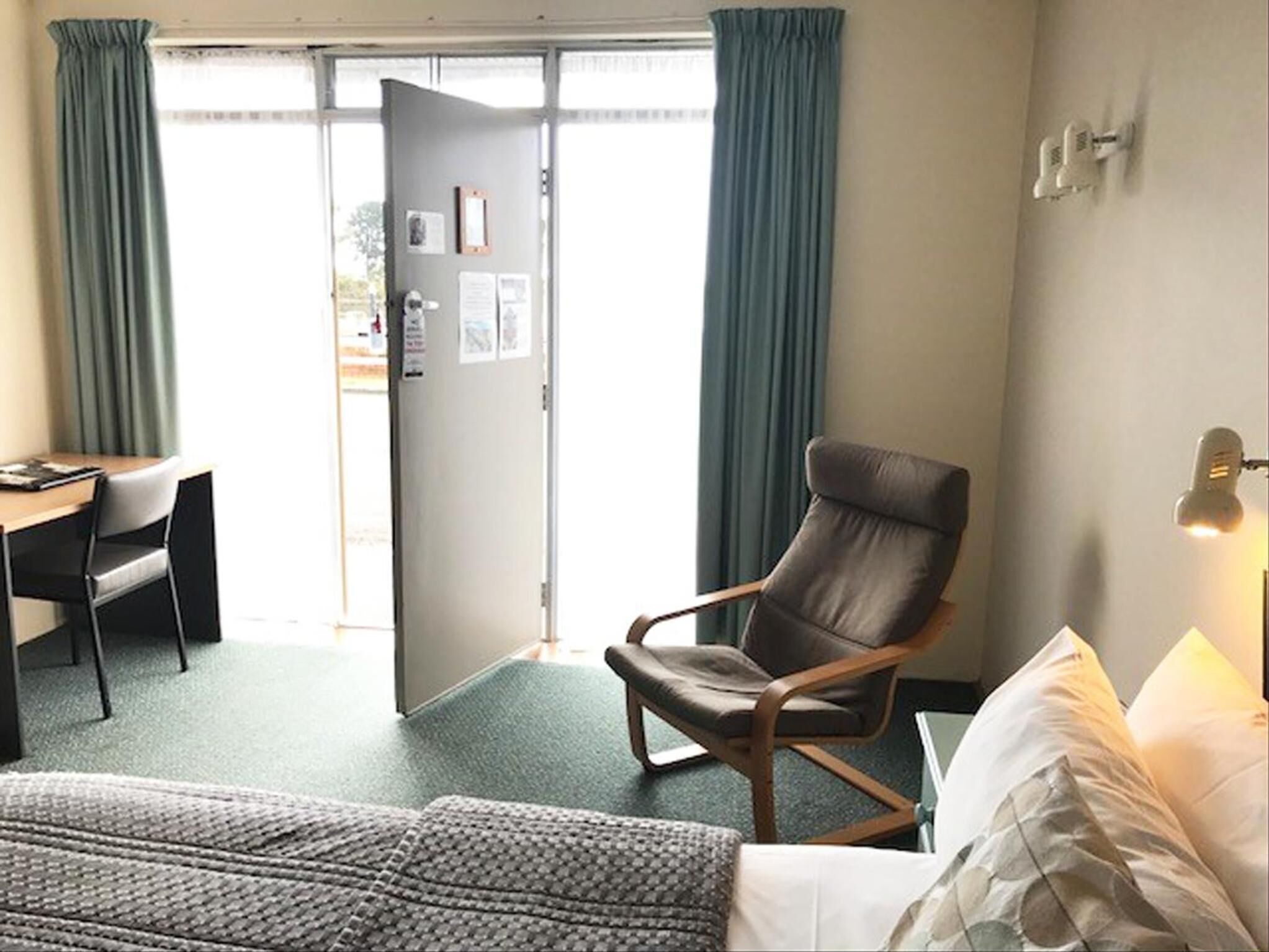 A hotel room with a bed, a desk, a chair, and a door leading to the outside.