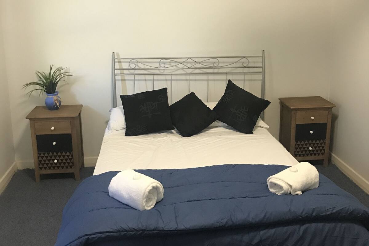 A neatly made bed with a blue bedspread and two white towels, flanked by two nightstands with decorative vases, a framed picture on the wall, and a simple, clean room layout.